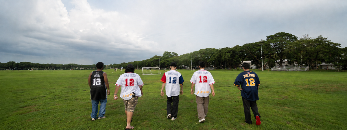 Five people walking away on a grassy sports field, each wearing a different sports jersey with the number 12 on the back. The jerseys include black, white, and navy blue options, with trees and a cloudy sky in the background.